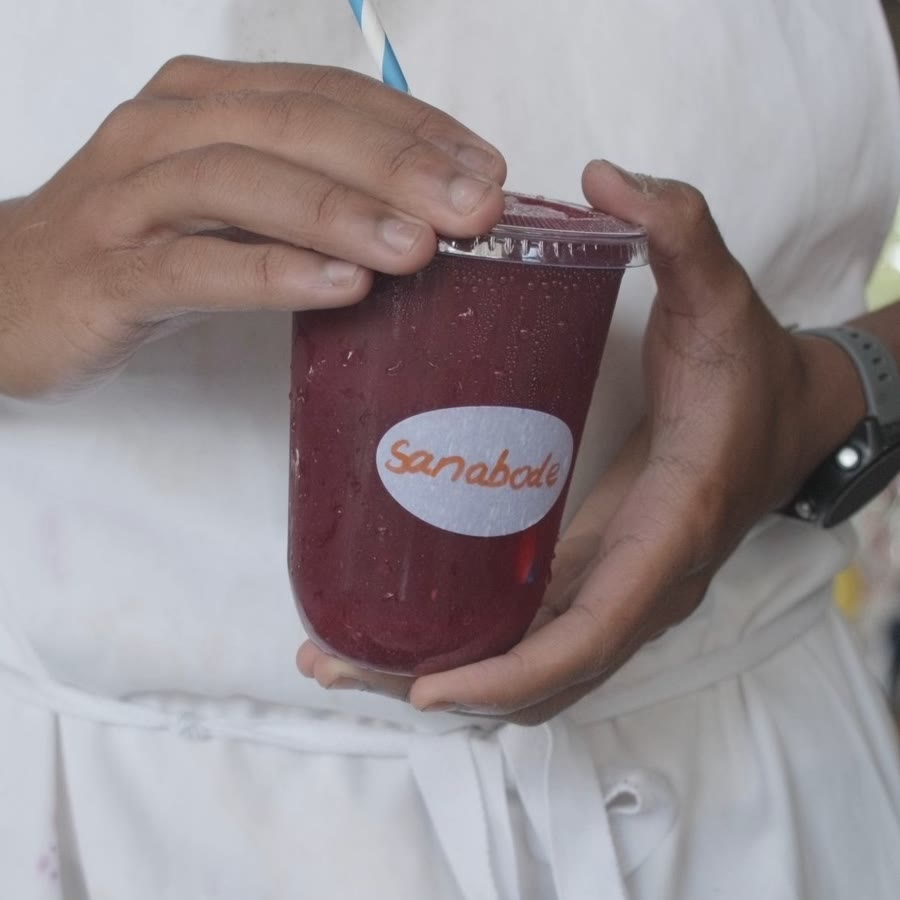 The Sanabode logo on orange beside a deep red fruit blend in a Sanabode cup